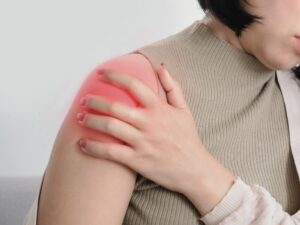 肩に強い痛みを感じている女性が、肩を押さえている様子。肩の部分が赤く強調されており、四十肩・五十肩などの痛みや炎症を象徴している画像。