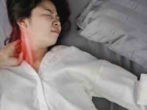 首のこりに悩み、痛みを感じて眠れない女性。首の後ろが赤く強調されており、慢性的な首のこりや痛みが原因で寝苦しい状態を表している。