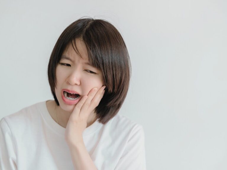 顎関節症の痛みにより顎を開けるのに苦労している女性。白い背景に白いTシャツを着用し、顔をしかめて顎に手を当てている様子。