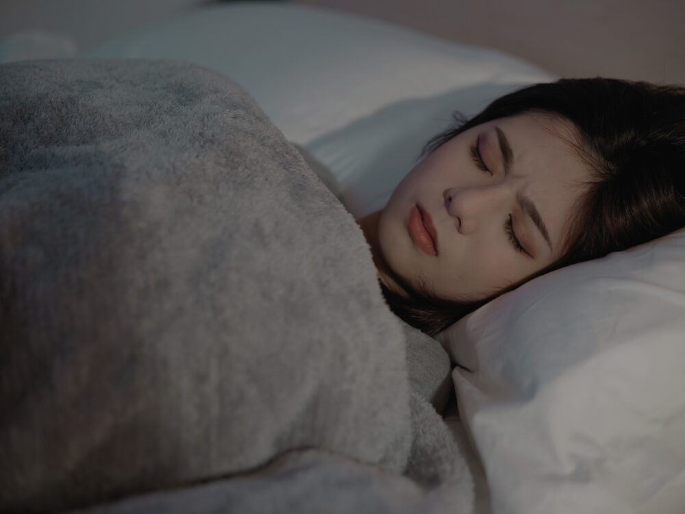 布団の中で眠れず、苦しそうな表情を浮かべている女性。快眠できない様子と疲労感が伝わるシーン。