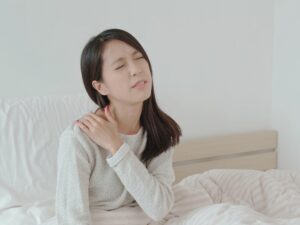 背中の痛みと慢性の肩こりで浅い眠りに悩まされ、疲労が解消されない様子の女性がベッドの上で肩を押さえながら痛みを感じている姿。