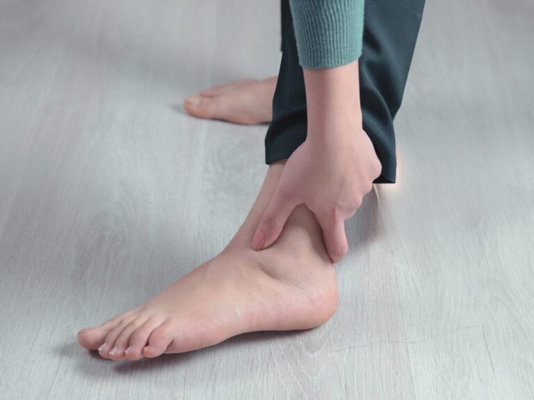 左足のくるぶし周辺を両手で押さえている女性の足元。体重をかけると足首に痛みがある様子を示している。