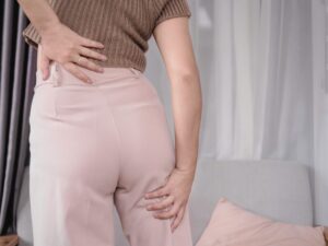 右のお尻から太もも、ふくらはぎにかけての痛みやしびれに悩む女性が、腰と脚を同時に手で押さえている様子。坐骨神経痛の症状を示している。