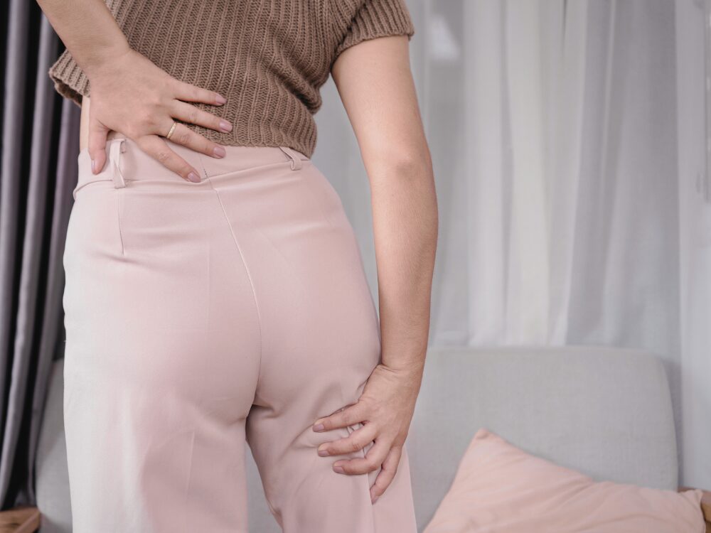 右のお尻から太もも、ふくらはぎにかけての痛みやしびれに悩む女性が、腰と脚を同時に手で押さえている様子。坐骨神経痛の症状を示している。