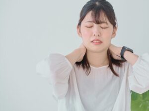 首の痛みで動かせずに辛そうな表情を浮かべる女性。両手で首の後ろを支え、目を閉じて耐えている様子。