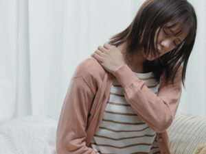 肩こりのつらさに目を閉じて耐える女性。左肩を手で押さえ、痛みや重だるさを感じている様子がうかがえる。