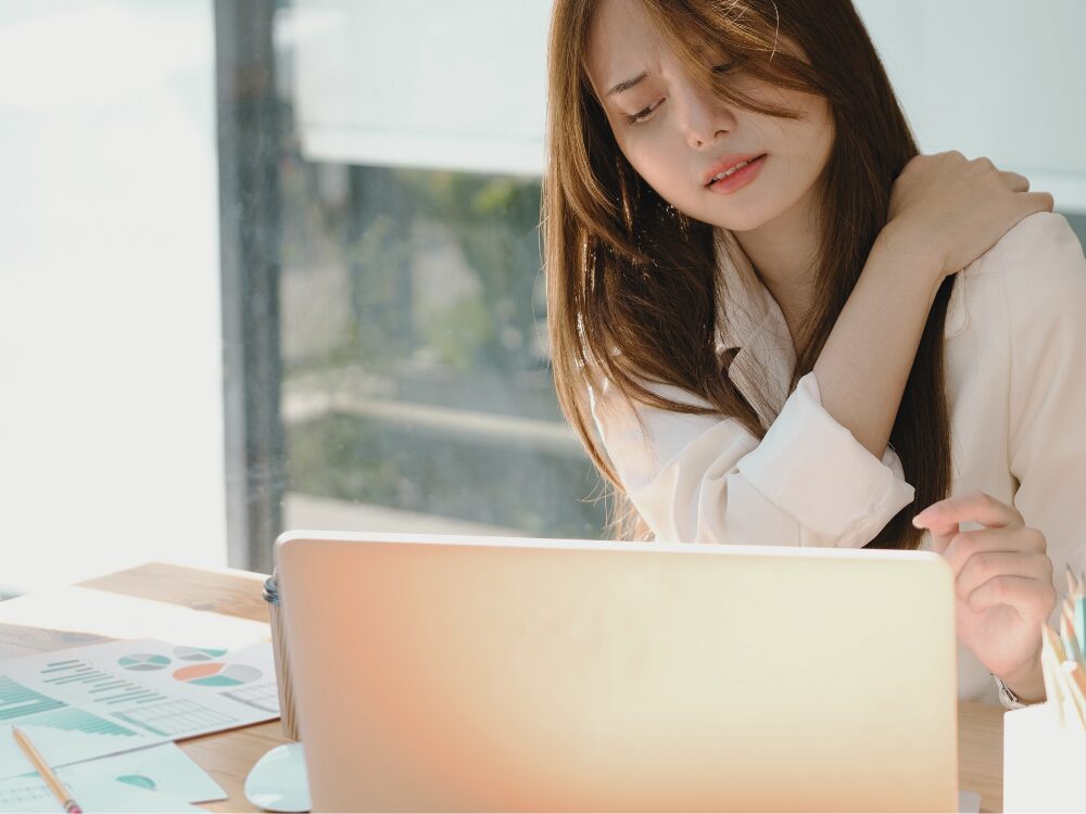 パソコン作業中に肩の痛みを感じ、右肩を押さえている女性。デスクに書類とノートパソコンがあり、長時間のデスクワークによる肩こりの様子を表している。